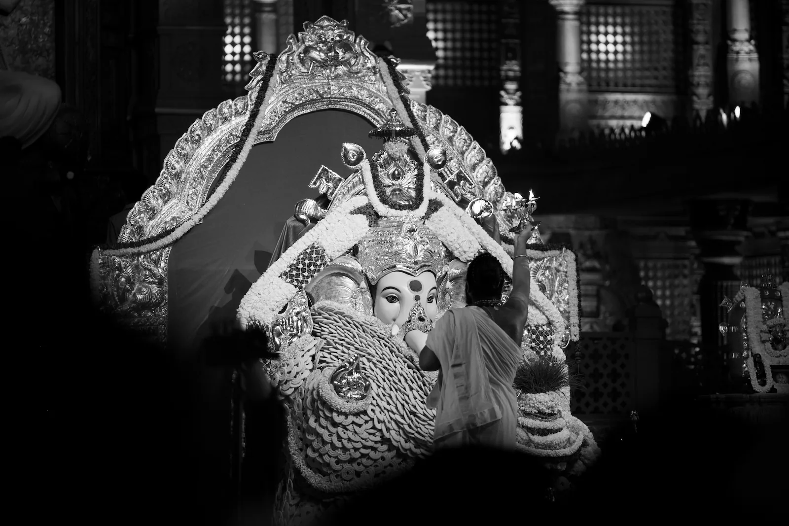 A woman in a saree holding up an aarti lamp to the Ganapathi idol, which sits under a silver arch hung with garlands