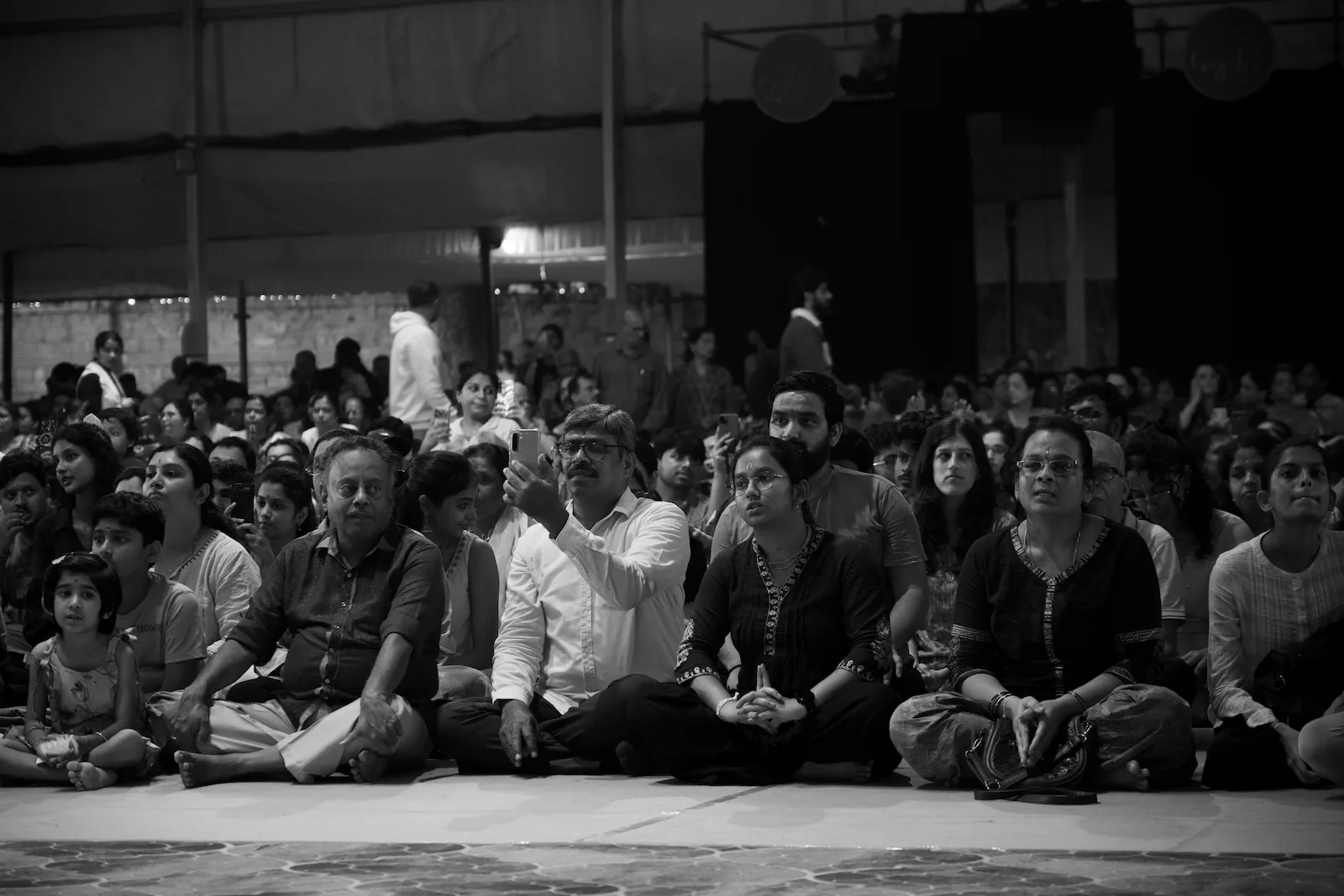 The audience sitting cross-legged on the floor, some with their hands folded and one man holding up his phone to record