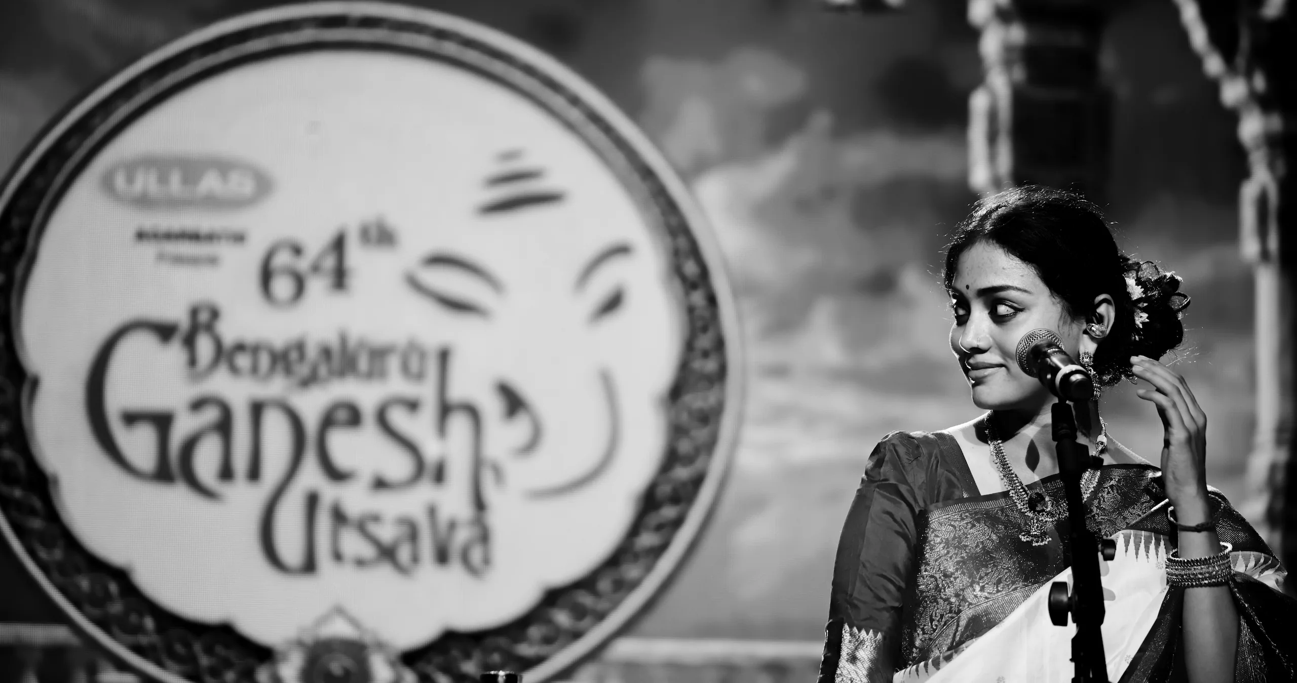 Sivasri Tejasvi Surya smiling at the microphone on stage, with the 64th Bengaluru Ganesh Utsava logo on the screen behind her