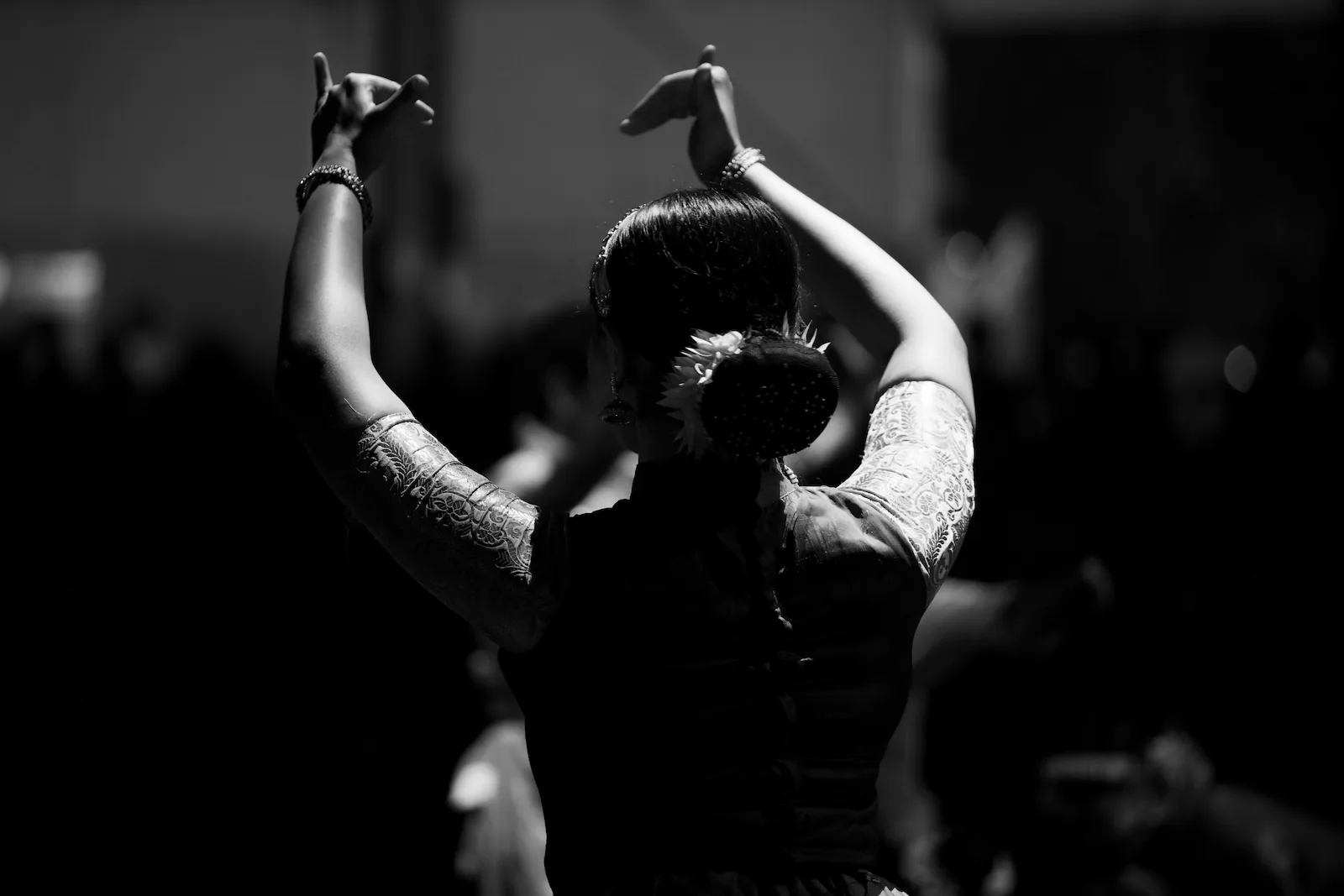 A dancer seen from behind with both arms raised and her hands in a mudra, flowers in her hair