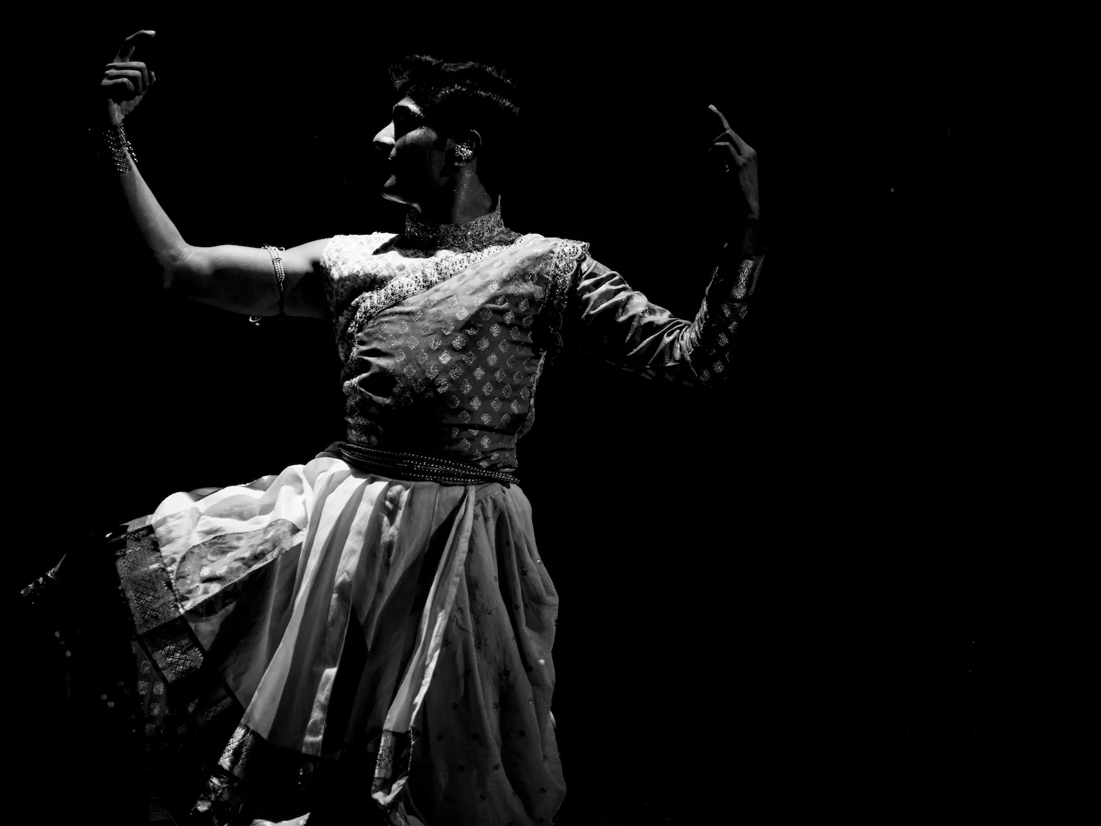A male dancer lit against a dark stage, both hands raised and the skirt of his costume flared out