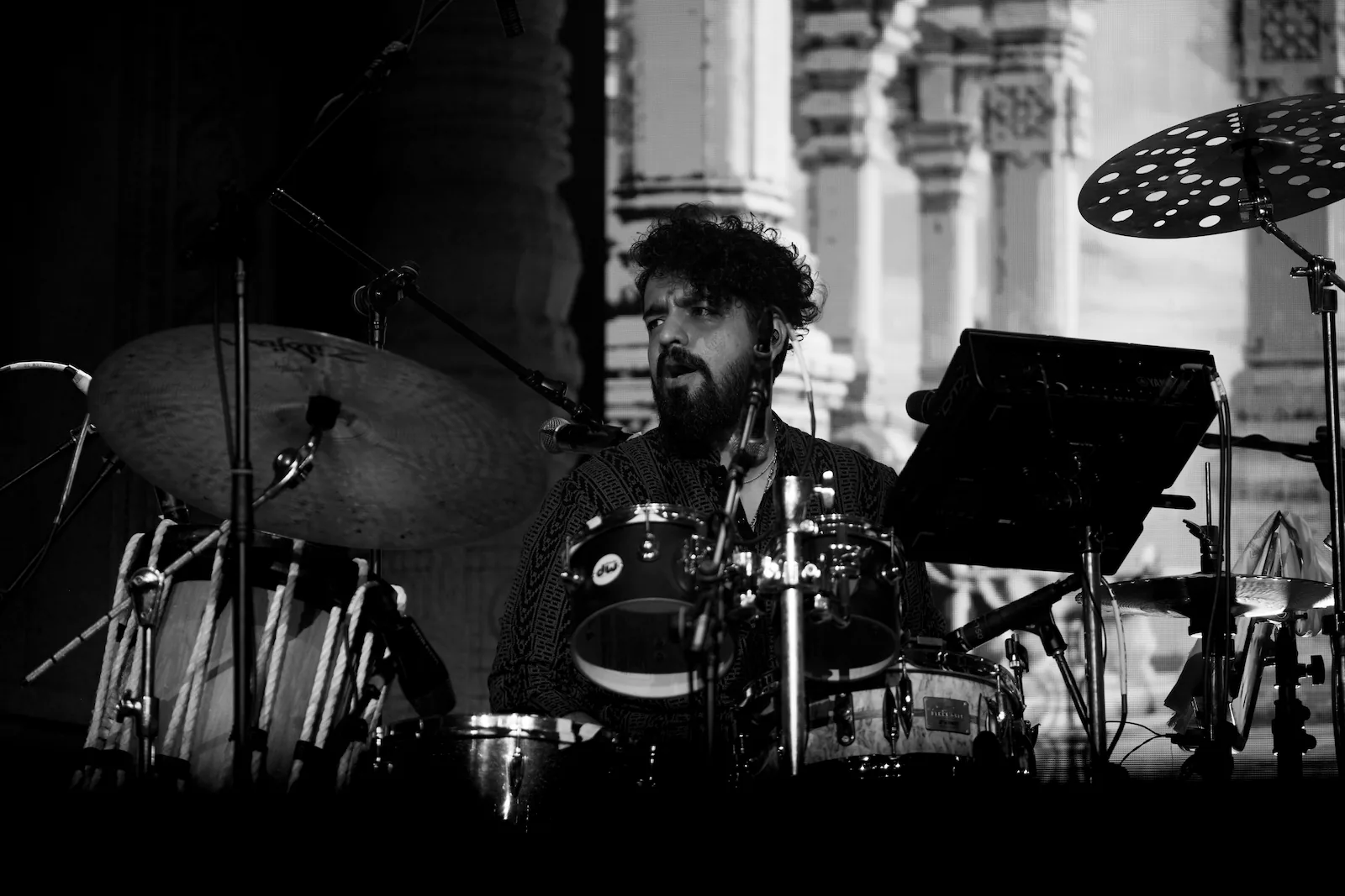A drummer with curly hair behind a drum kit and cymbals, with a rope-tuned drum beside him