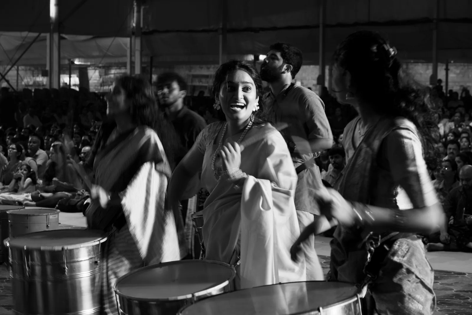 A drummer in a saree laughing mid-beat, the drummers on either side of her blurred by their movement, and the audience sitting behind them