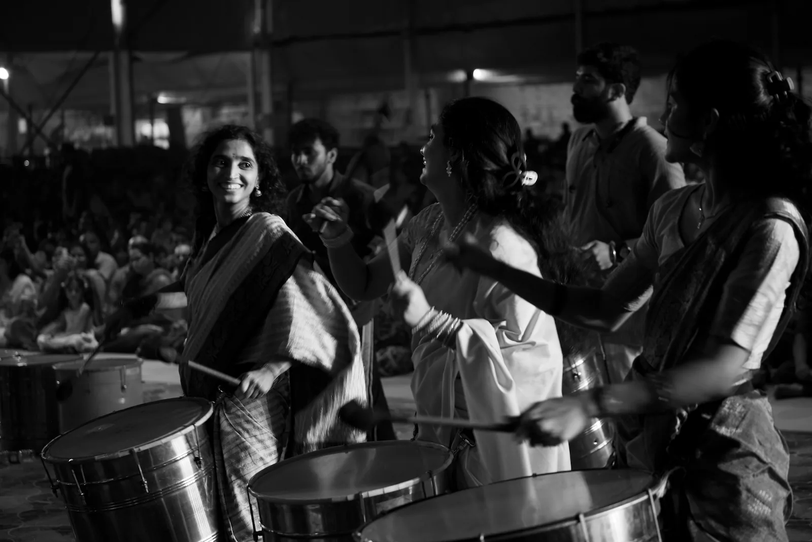 Women in sarees playing big steel drums with sticks, one of them smiling, with men in kurtas behind them and the audience sitting on the floor