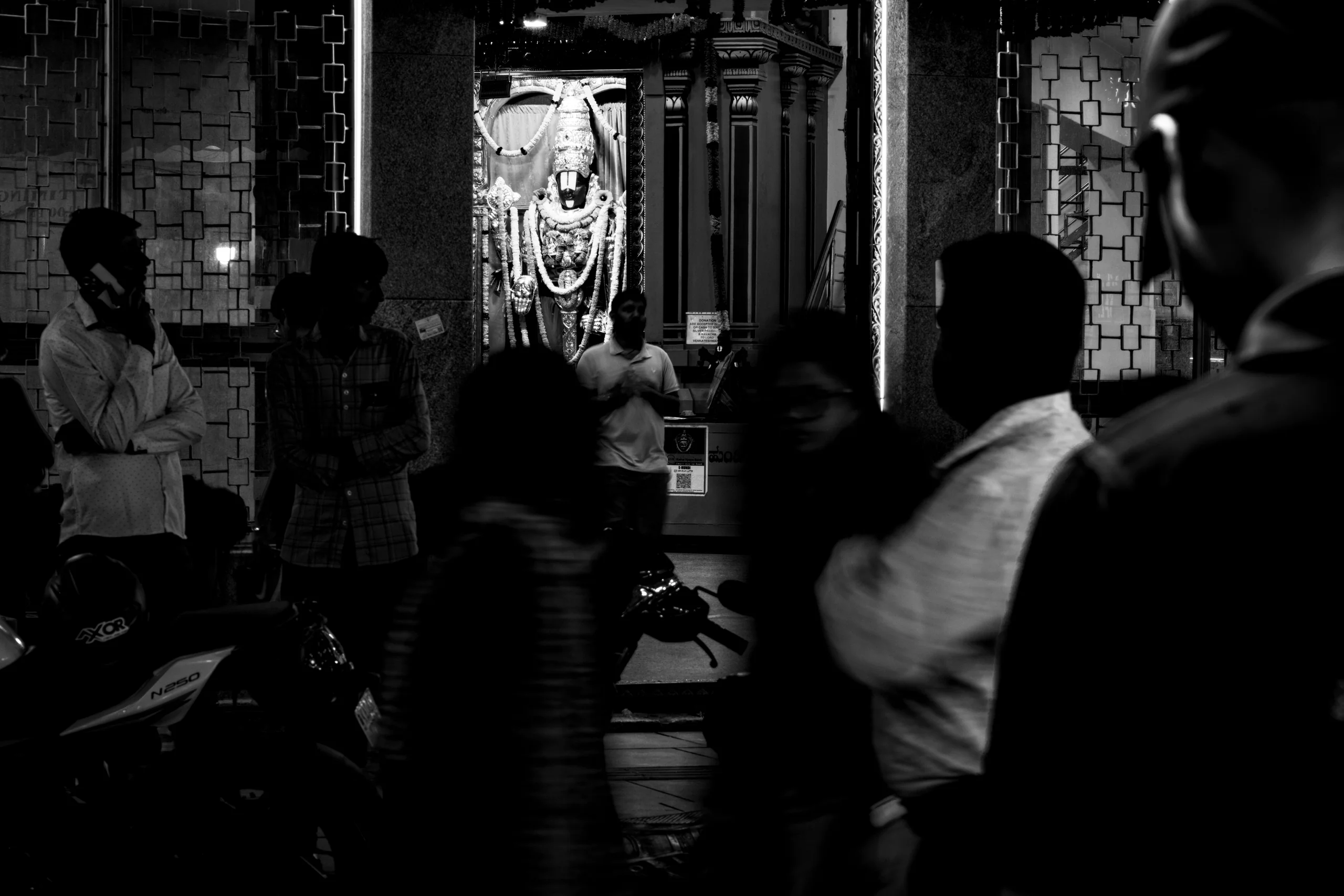 A roadside shrine glows behind the everyday traffic of people and bikes, with the deity steady at the centre. Everything else is in motion around it - the priest's gaze, the stranger on her phone, and the man crossing the frame.