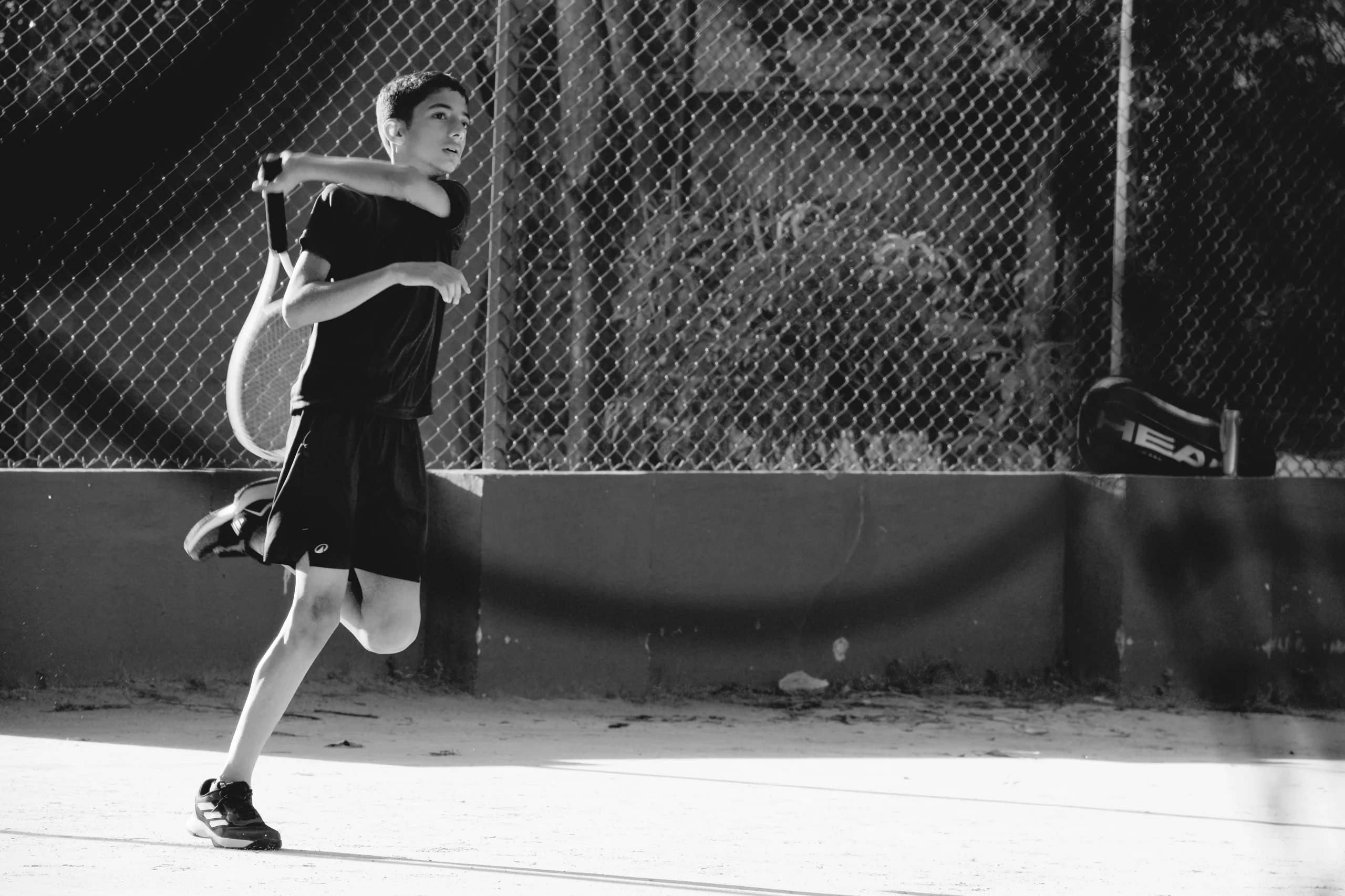 This is the end of a forehand, held at the moment the whole body is committed to a shot that's already gone, with the eyes tracking the ball down the court. The racquet is already behind the shoulder and the back foot still off the ground, in early light, with the chain-link and the long shadow of the wall behind.