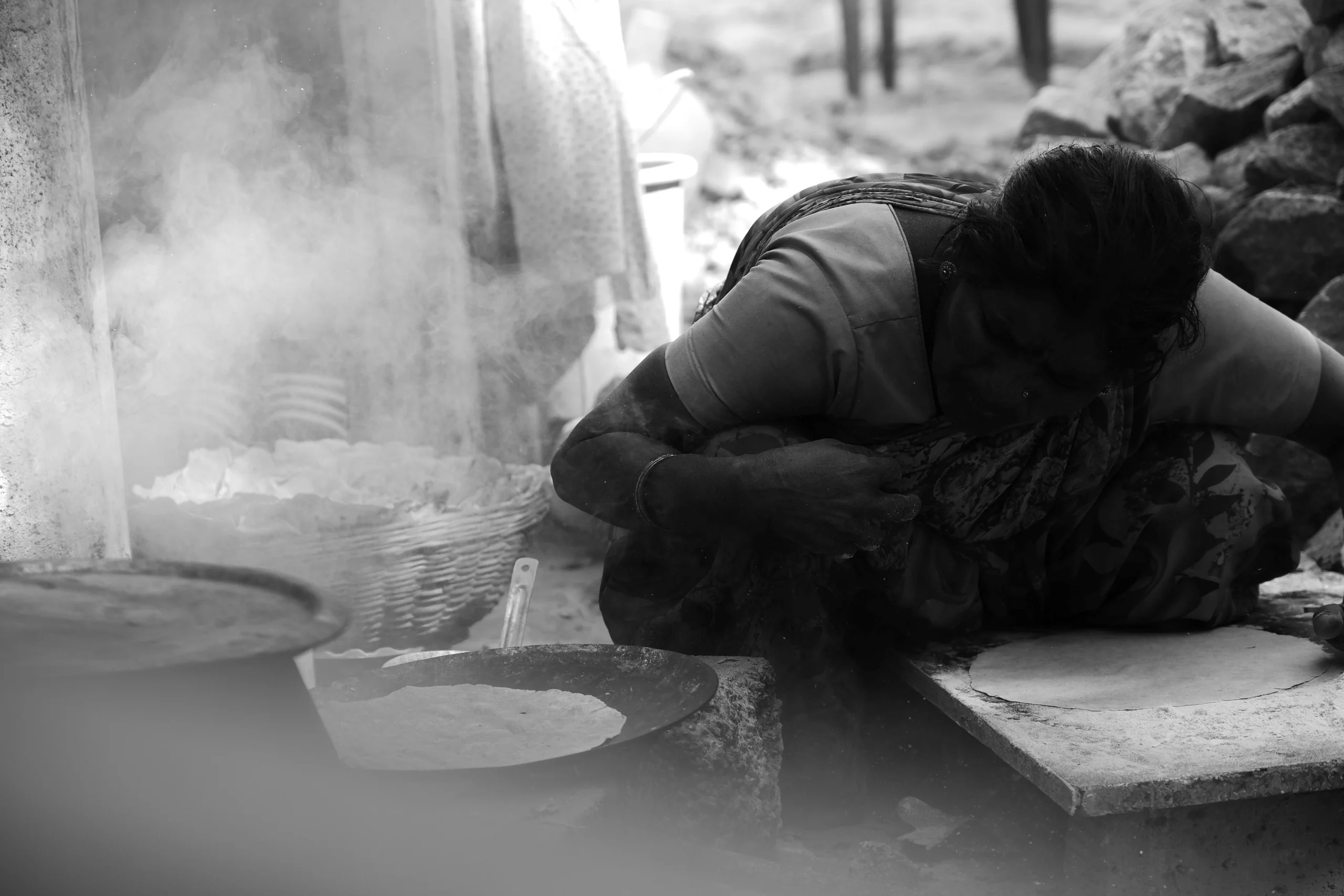 The day's first heat comes from someone else's labour - a woman at the rolling stone with dough in one hand. Steam rises from the tava, with a basket of finished roti waiting in the morning light.