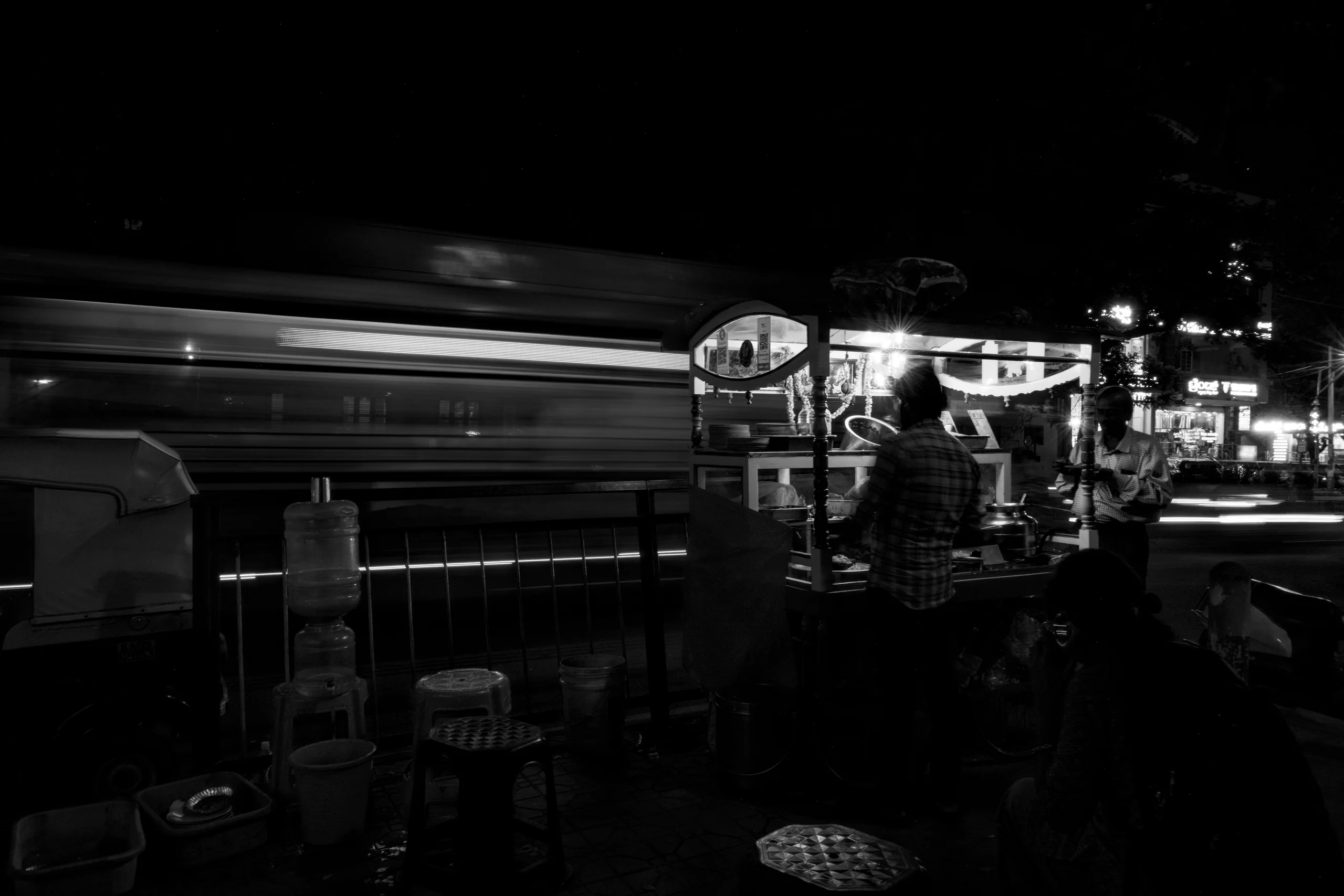 A street stall lit against a quiet road is a small, bright place of work, with the city slowing down around it. The vendors are finishing up for the night while the traffic on the left leaves trails in the long exposure.