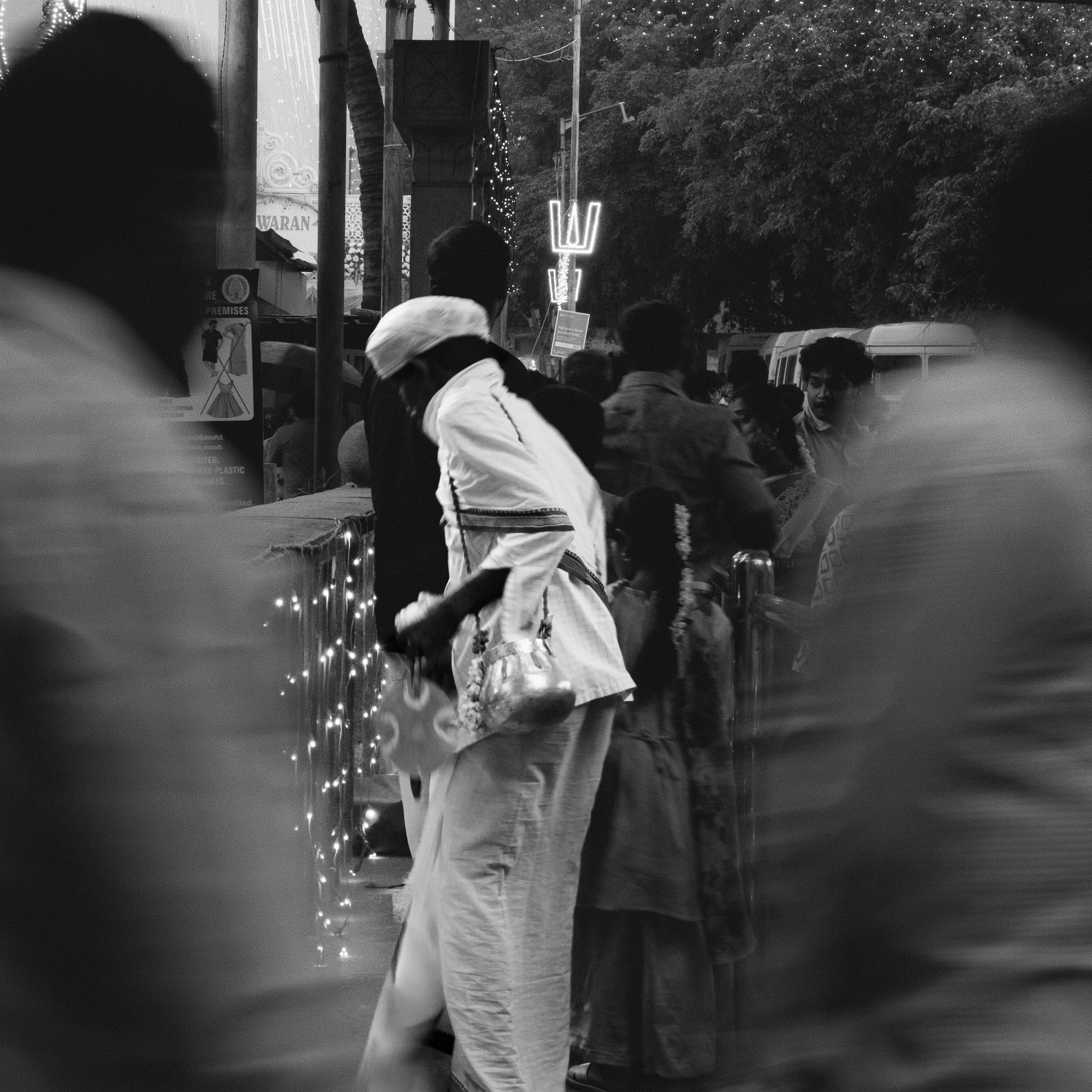 One man stands still while a temple queue moves past at a fifteenth of a second, with everyone in the frame smeared except him. Fairy lights hang down the barrier and a trishula is picked out in bulbs above.