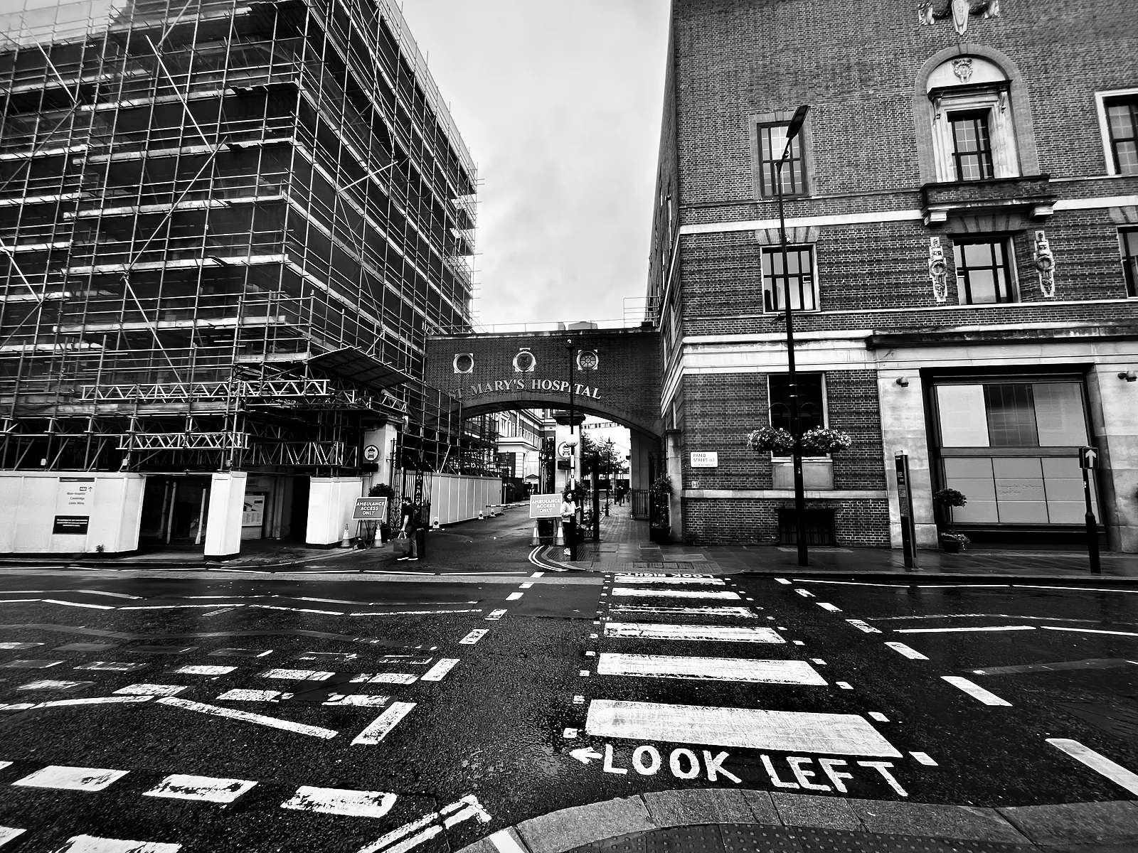 A wet road crossing painted LOOK LEFT beside St Mary's Hospital in Paddington, with scaffolding on the building opposite