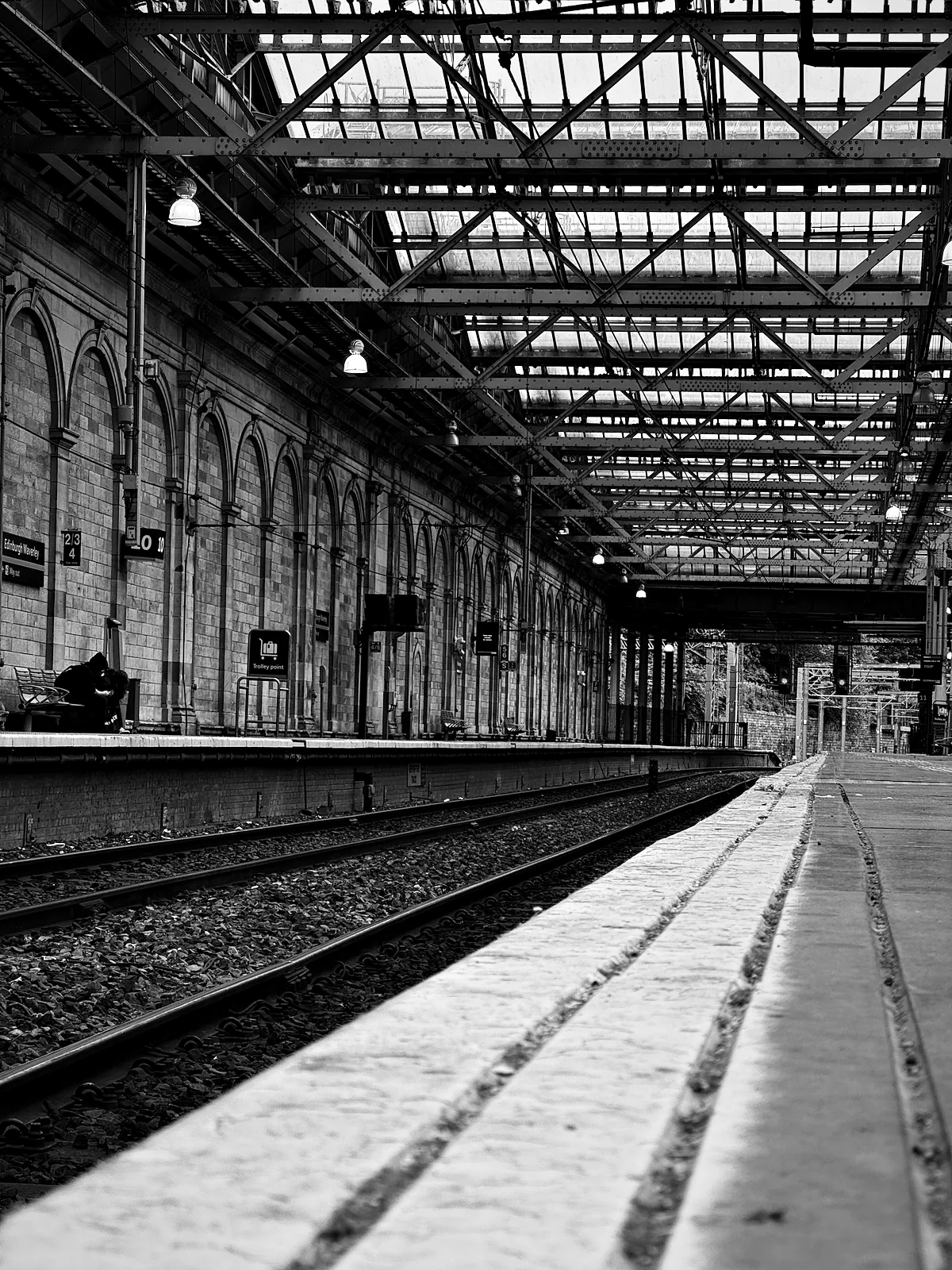 An empty platform under the iron and glass roof of Edinburgh Waverley, with one person sitting across the tracks