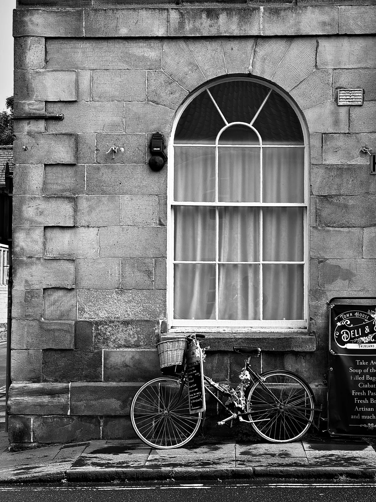 A bicycle with a basket and flowers tied along its frame, parked under an arched window in Falkland