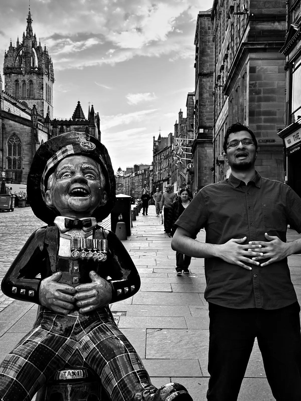 Viren laughing with his hands on his belly beside a laughing statue of Oor Wullie in tartan on the Royal Mile, with St Giles' crown spire behind