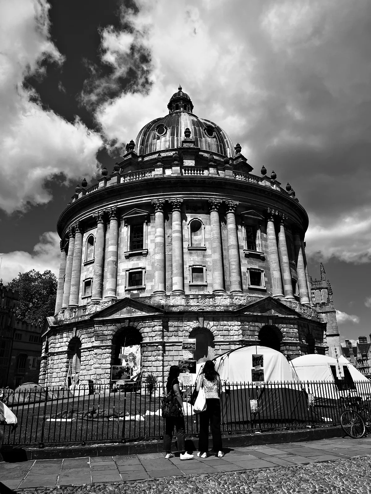 The Radcliffe Camera in Oxford under big clouds, with protest tents on the lawn behind its railings