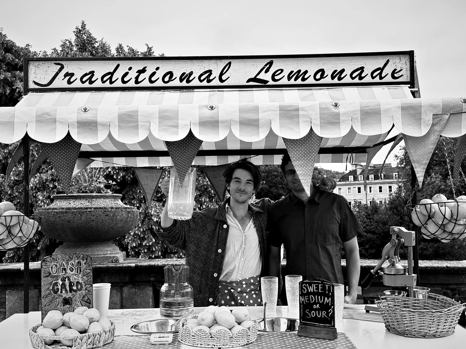A Traditional Lemonade stall near Pulteney Bridge in Bath, the stallholder raising a jug with Viren beside him behind the counter, and a chalkboard asking sweet, medium or sour